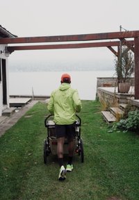 En person i en knallgrønn jakke skyver en barnevogn langs en gresskledd sti, omgitt av trebygninger med en innsjø i bakgrunnen.