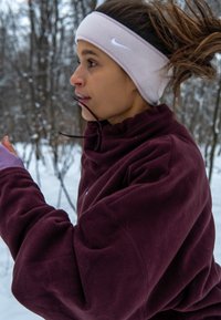Jeune femme faisant du jogging dans une forêt enneigée, portant un polaire bordeaux et un bandeau Nike beige, sa queue de cheval oscillant derrière elle.