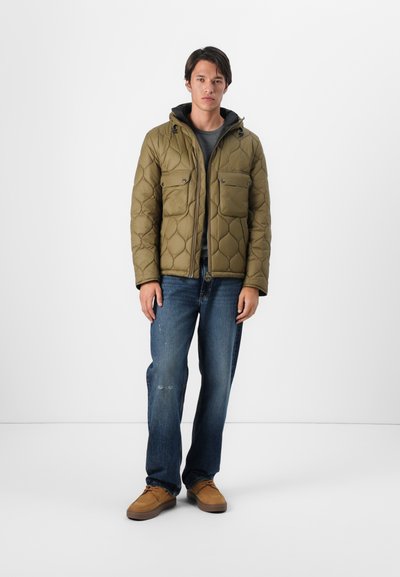 Chaqueta tipo puffer acolchada en color verde oliva con capucha, que presenta dos bolsillos en el pecho y un tejido texturizado, combinada con jeans azules y zapatos marrones.