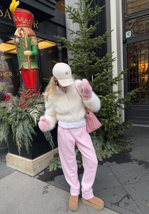Chaqueta de piel sintética blanca con detalles en rosa, pantalones deportivos rosa pastel, botas beige con plataforma y una gorra clara. Decoración de ramas navideñas al fondo.