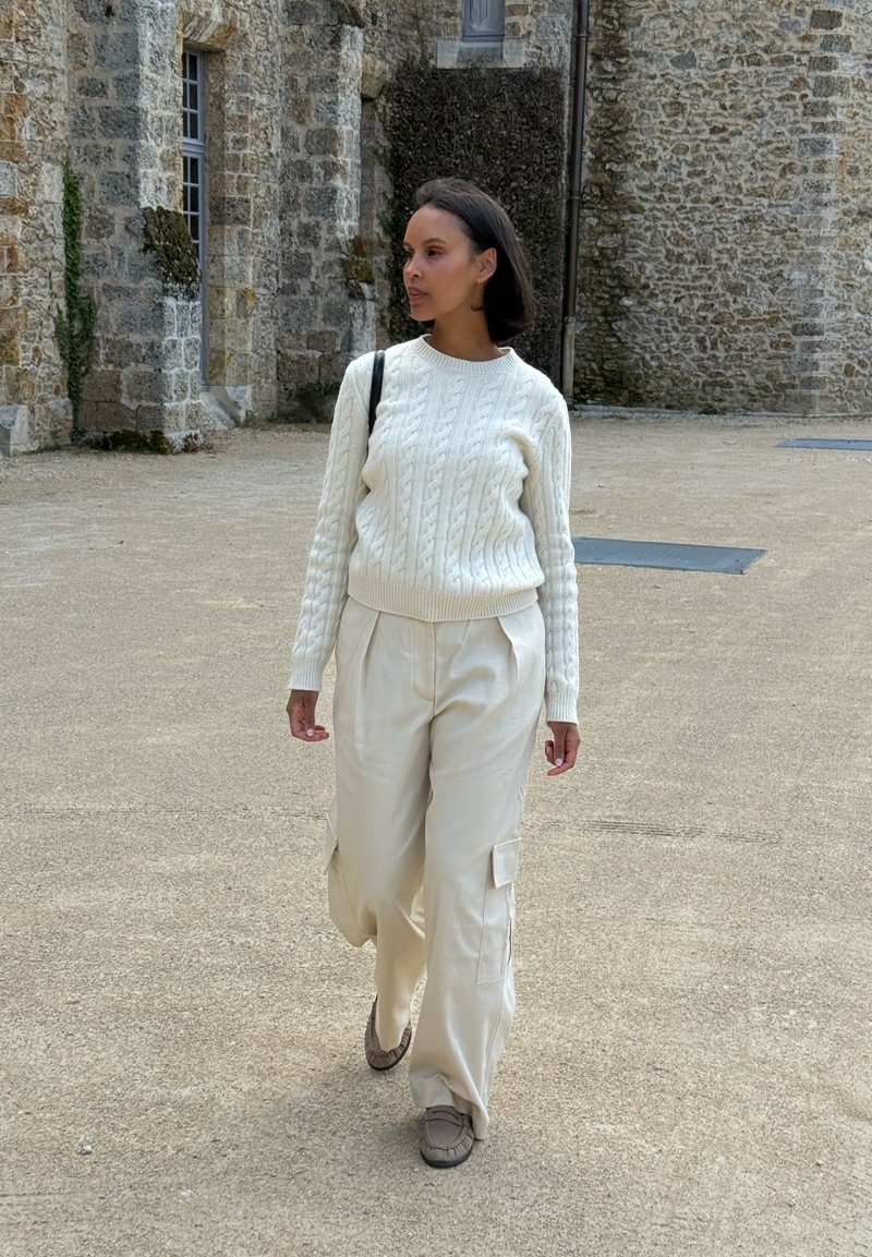 Femme marchant à l'extérieur près d'un bâtiment en pierre, portant un pull en maille torsadée blanc, un pantalon cargo crème et portant un petit sac noir à l'épaule.
