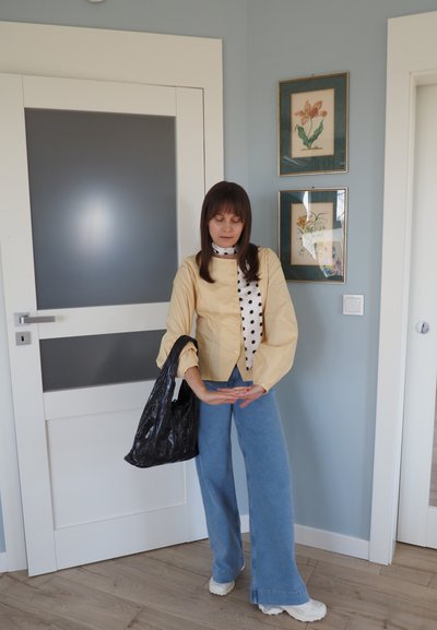 Mujer en interiores con blusa amarilla, pañuelo de lunares, vaqueros azules, zapatillas blancas, sosteniendo un bolso negro, de pie junto a una puerta blanca con arte floral enmarcado en la pared.