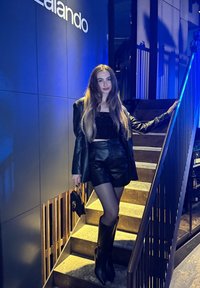 Black leather blazer and shorts, sequined top, sheer tights, knee-high black boots. Standing on stairs with wooden accents. Blue lighting.