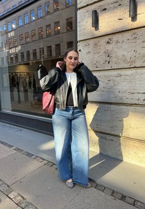 Joven mujer se arregla el cabello mientras está de pie en la acera de la ciudad junto a una pared de piedra, lleva chaqueta de cuero negra, camiseta blanca, jeans de pierna ancha y lleva un bolso rojo.