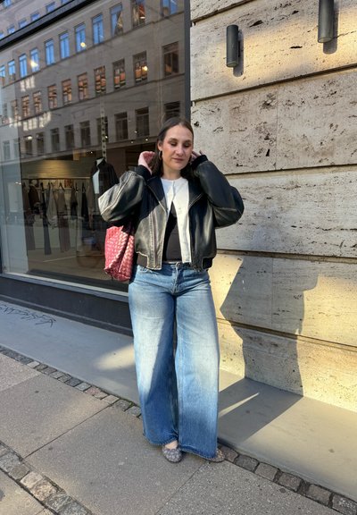Joven mujer se arregla el cabello mientras está de pie en la acera de la ciudad junto a una pared de piedra, lleva chaqueta de cuero negra, camiseta blanca, jeans de pierna ancha y lleva un bolso rojo.
