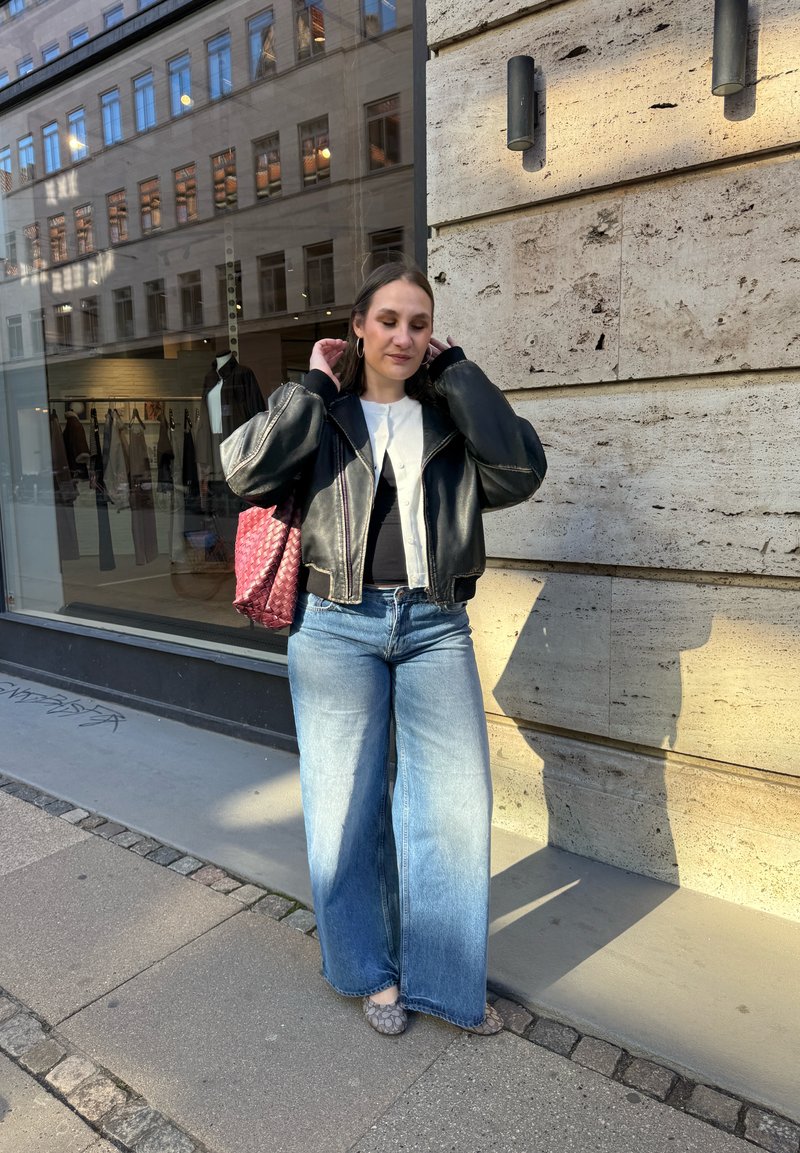 Junge Frau richtet ihre Haare, steht auf dem Bürgersteig einer Stadt neben einer Steinmauer, trägt eine schwarze Lederjacke, ein weißes Top, weit geschnittene Jeans und trägt eine rote Tragetasche.