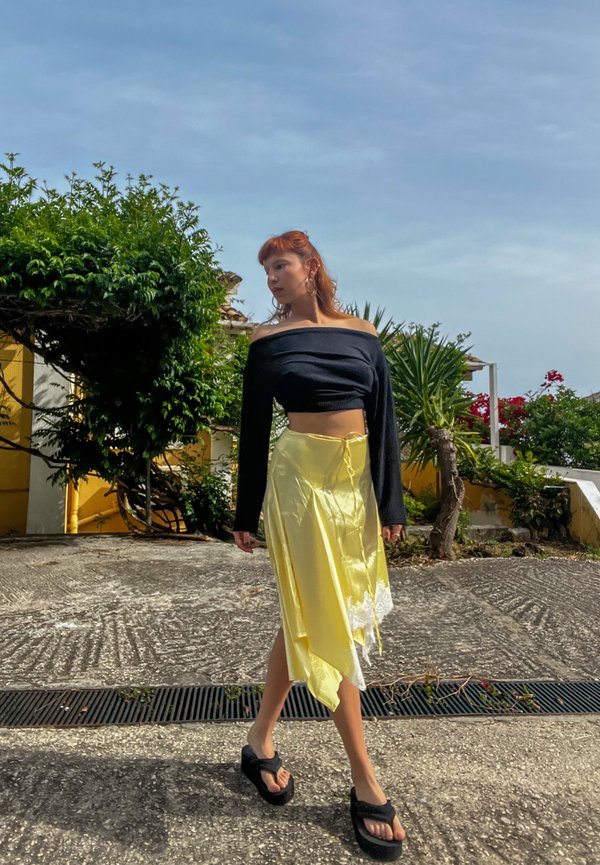 Frau mit roten Haaren, die ein schwarzes schulterfreies Oberteil, einen gelben Rock und schwarze Plateau-Sandalen trägt, geht draußen in der Nähe von Grünanlagen und Gebäuden spazieren.