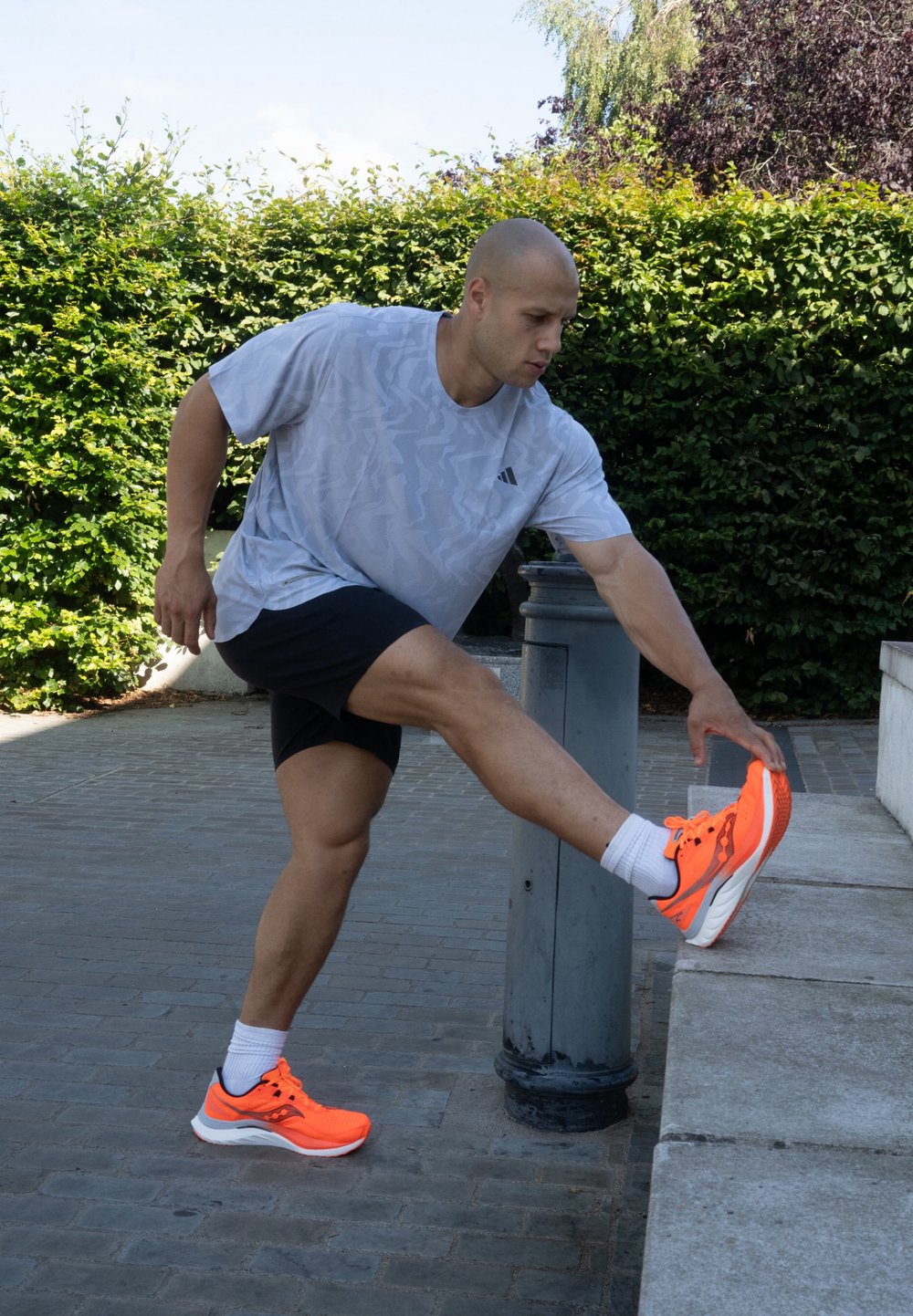 Sportliche Schuhe in leuchtendem Neonorange mit einer strukturierten Oberseite und gepolsterter Sohle. Modell in grauem gemustertem Shirt und schwarzen Shorts, das/der sein/ihr Bein dehnt.