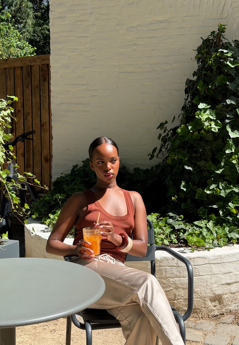 Jeune femme avec un débardeur marron et un pantalon beige assise à l'extérieur, tenant un verre de boisson orange près d'un mur vert couvert de lierre.