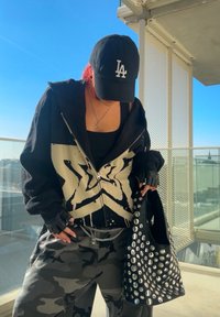 Hoodie preto com design de estrela branca, top preto, calças de camuflagem e uma grande mala preta com tachas prateadas. Boné preto com o logótipo "LA".