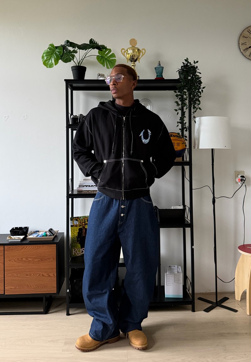 Sweat à capuche noir zippé avec coutures blanches et logo, associé à un pantalon en denim ample et des sabots couleur sable. Environnement intérieur avec des plantes et des étagères.