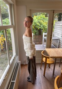 Witachtige, off-shoulder trui met lange mouwen, gecombineerd met oversized grijze broek, staand naast een raam in een houten interieur.