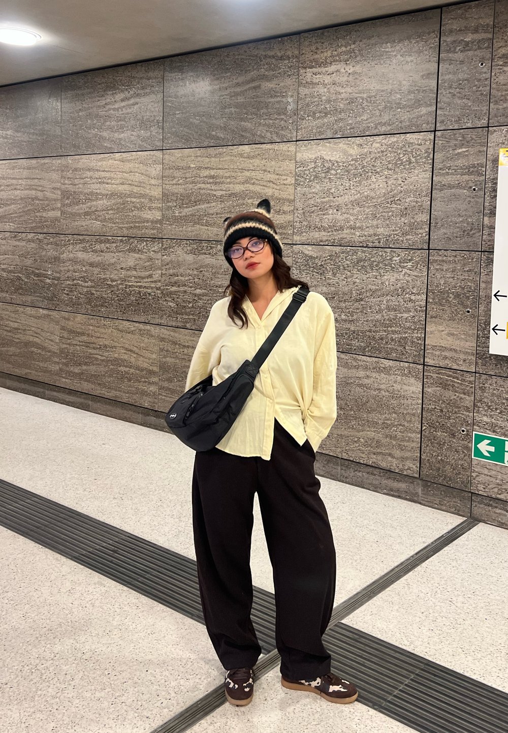 Jeune personne portant des lunettes, un bonnet, une chemise jaune, un sac noir et un pantalon large noir debout dans une station de métro aux murs en pierre texturée.
