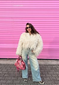 Bílá chlupatá krátká bunda, světle modré široké džíny, černé sluneční brýle a růžová kabelka, pózující před růžovou pruhovanou stěnou.