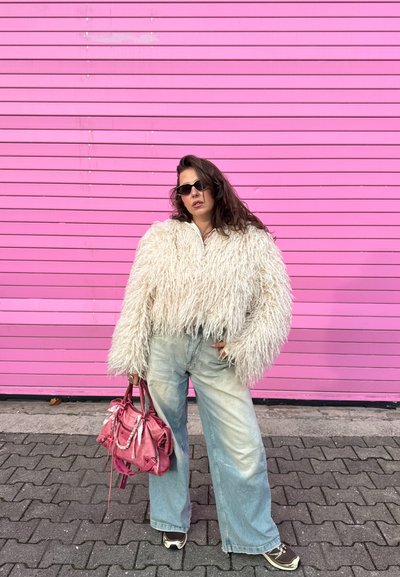 Chaqueta corta blanca y peluda, vaqueros anchos de pierna en azul claro, gafas de sol negras y un bolso rosa, posando frente a una pared con rayas rosas.
