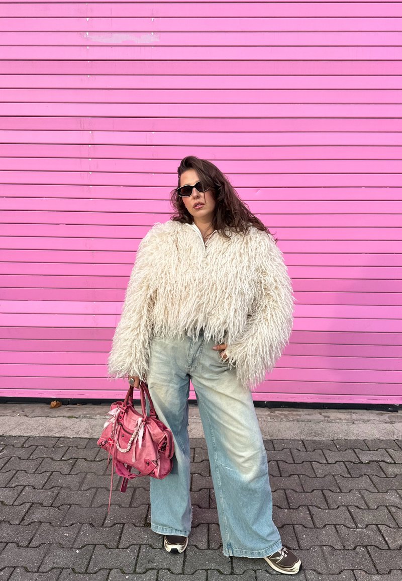 Giacca corta bianca pelosa, jeans azzurri a gamba larga, occhiali da sole neri e borsa rosa, posati contro un muro a righe rosa.