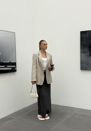 Bežinis lininis švarkas virš baltos trumpinuko, juoda satino sijonas ir balti platforminiai sandalai. Laiko mažą baltą rankinę. Minimalistinė galerijos fono aplinka.