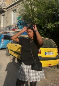 Μαύρο μπλουζάκι, καρό φούστα με ραντάρ, μαύρη τσάντα με λογότυπο και καλσόν. Κίτρινο αυτοκίνητο και γκράφιτι στο φόντο. Ηλιόλουστη εξωτερική ατμόσφαιρα.