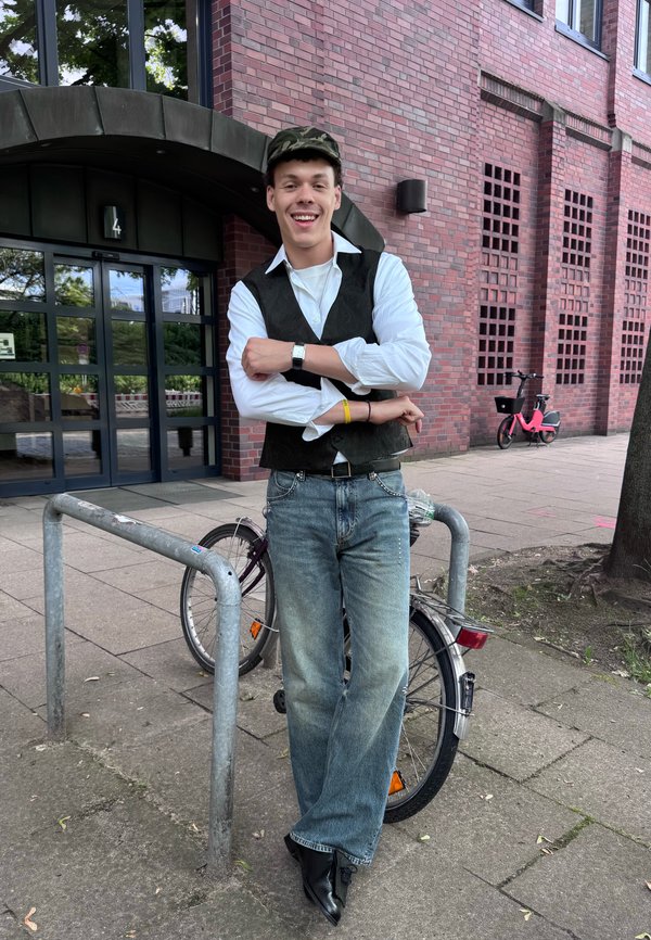 Młody mężczyzna w casualowym ubraniu i czapce opiera się o zaparkowany rower przed ceglaną budowlą ze szklanymi drzwiami, uśmiechając się ze skrzyżowanymi rękami.