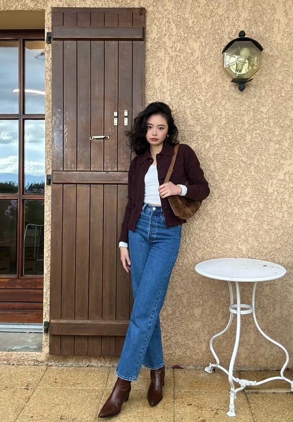 Une femme portant une veste bordeaux, un haut blanc, un jean et des bottes brunes pose à côté d’un volet en bois et d’une petite table blanche.