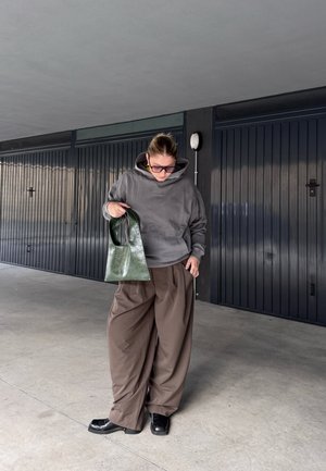 Sudadera gris con capucha, pantalones sueltos marrones, zapatos negros, sosteniendo una bolsa verde brillante con un asa redonda, textura suave, entorno urbano.