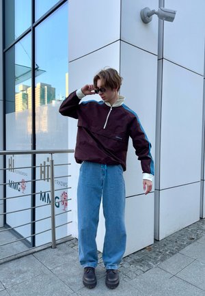 Chaqueta de media cremallera burdeos con mangas de rayas azules, combinada con unos jeans sueltos de color azul claro y botas negras gruesas. Lleva gafas reflectantes.