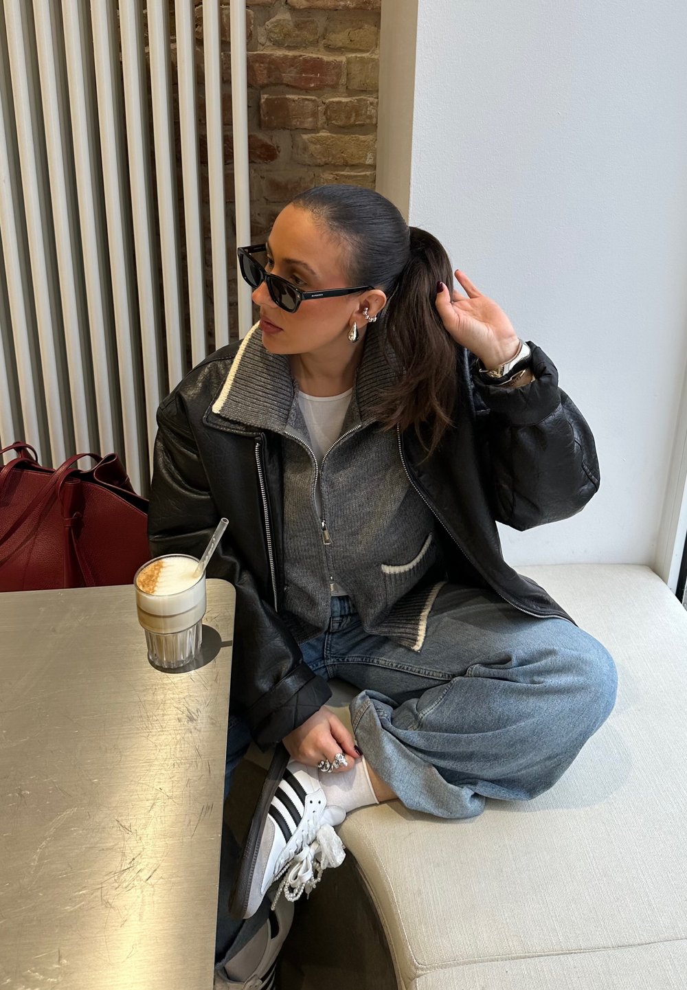 Μαύρο δερμάτινο μπουφάν με λευκό γιακά, γκρι πουλόβερ, ανοιχτό μπλε τζιν και λευκά sneakers. Ποτήρι με κα�φέ με στρώματα πάνω σε μεταλλικό τραπέζι.