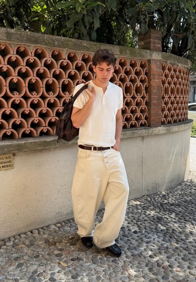 Joven con camisa blanca y pantalones beige holgados está parado en una calle adoquinada cerca de una pared de ladrillo y terracota, sosteniendo una mochila negra sobre el hombro.