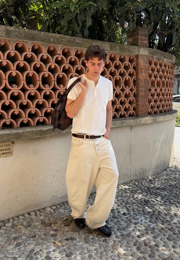 Giovane uomo con camicia bianca e pantaloni beige larghi in piedi su una strada di ciottoli vicino a un muro di mattoni e terracotta, con uno zaino nero sulla spalla.