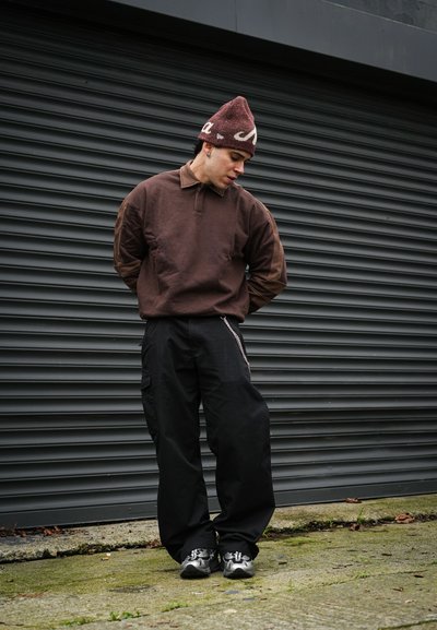 Joven vestido con un suéter marrón, gorro burdeos, pantalones anchos negros y zapatillas, de pie con las manos detrás de la espalda frente a una persiana metálica negra.