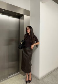 Brun læder midi-nederdel med en subtil glans, parret med en tætsiddende mørk top og spidse hæle. Sort håndtaske med guldfarvede detaljer.