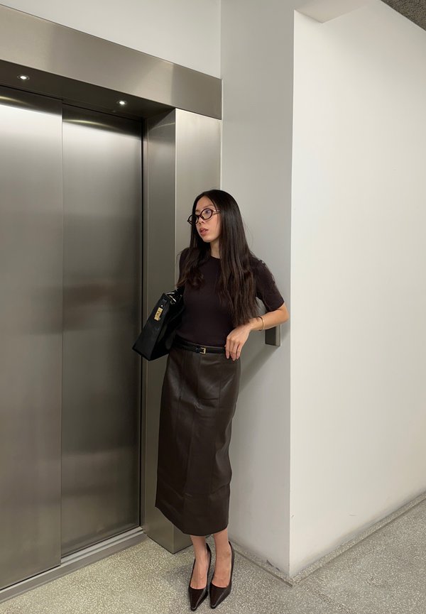 Brun læder midi-nederdel med en subtil glans, parret med en tætsiddende mørk top og spidse hæle. Sort håndtaske med guldfarvede detaljer.