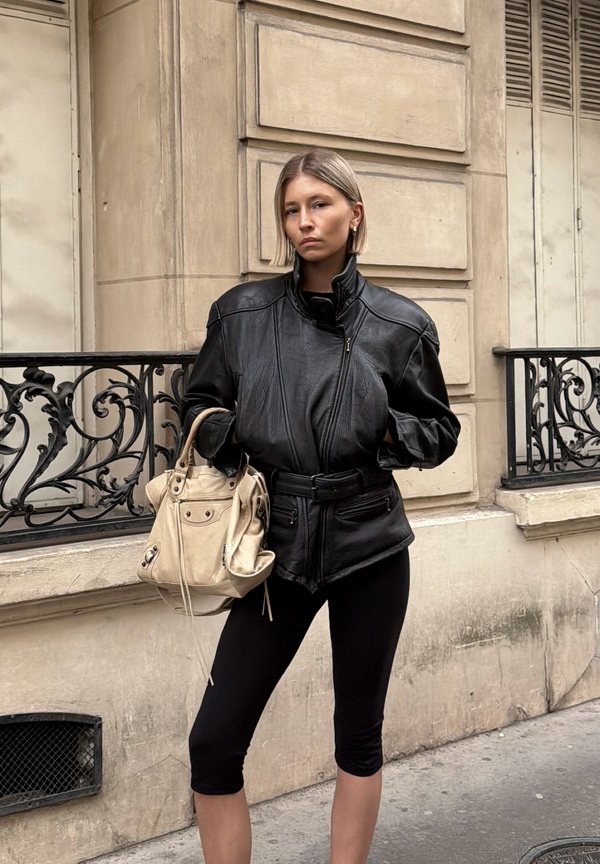 Młoda kobieta w czarnej skórzanej kurtce i legginsach stoi na ulicy miasta, trzymając beżową torebkę na tle kamiennego budynku.