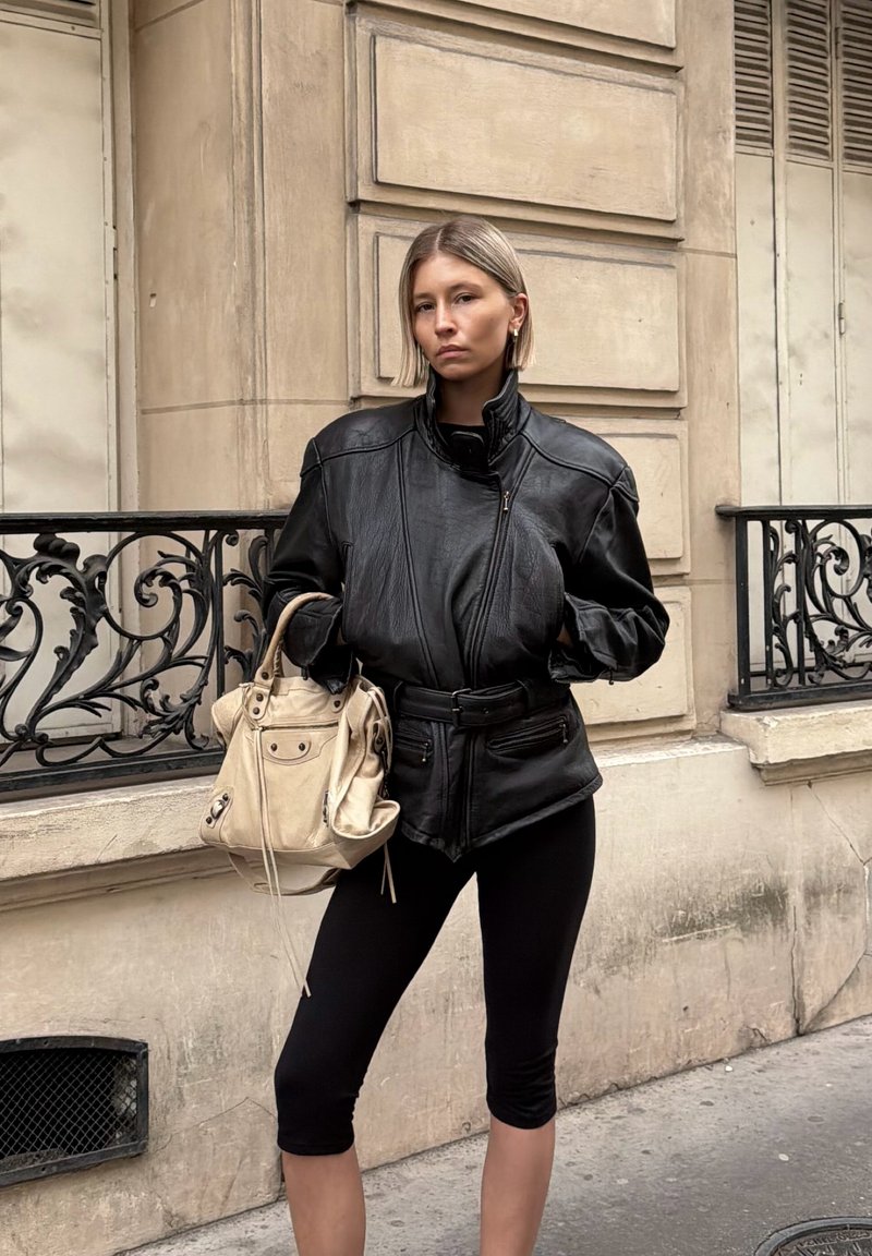 Junge Frau in schwarzer Lederjacke und Leggings steht in der Stadtstraße und hält eine beige Handtasche vor einem steinernen Gebäude.