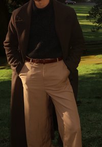 Homme portant un manteau marron foncé, un pull noir, un pantalon beige avec une ceinture tressée marron, debout dehors sur l'herbe les mains dans les poches.