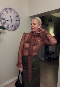 Bruine kanten blouse met gerimpelde kraag en veterdetail, gecombineerd met een aansluitende olijfgroene leren rok. Matte texturen en ingewikkelde patronen zichtbaar.