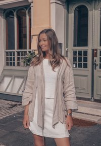 Ανοιχτό μπεζ λινό κάρντιγκ πάνω από λευκή υφαντή μπλούζα και λευκή φούστα, με χαλαρούς μανίκους και άνετο σχέδιο. Φόντο με πράσινη πόρτα.