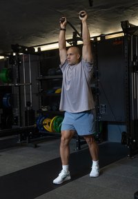 Uma pessoa vestindo uma t-shirt cinza e calções azuis claros realiza um exercício de cabo com cordas, destacando um ambiente de ginásio e equipamento de fitness.