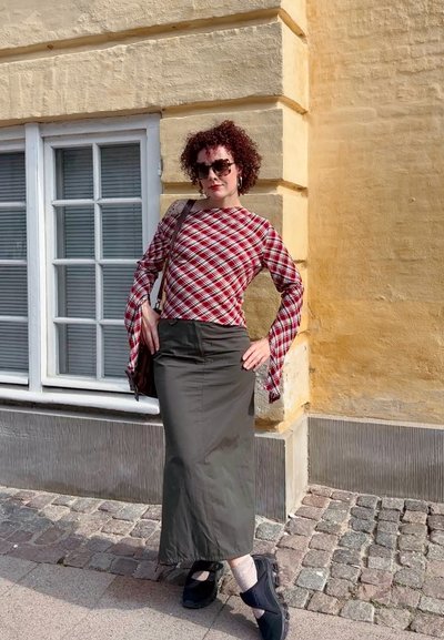 Blusa de manga larga a cuadros en rojo y blanco con mangas acampanadas, falda midi verde, calzado negro, gafas de sol, de pie frente a una pared amarilla.