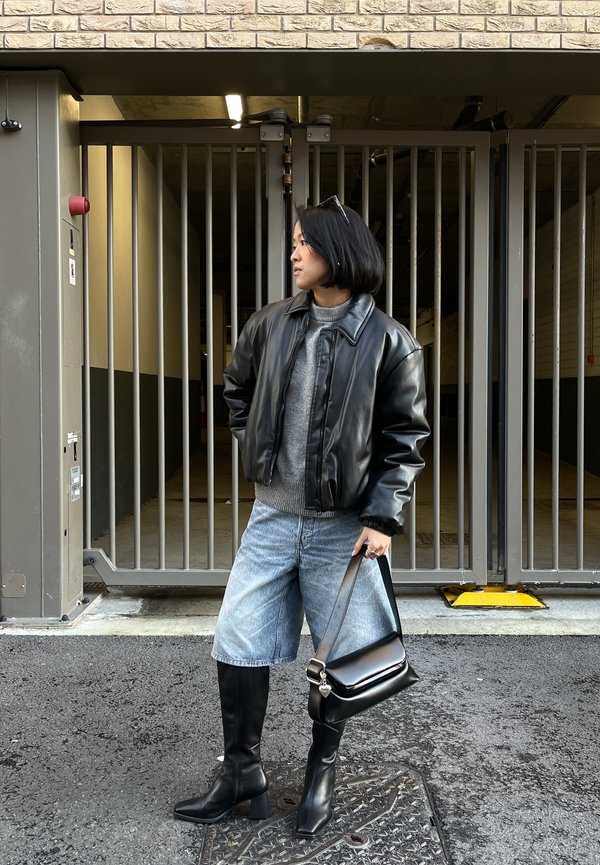 Frau in schwarzer Lederjacke, grauem Pullover, Jeansshorts und schwarzen Stiefeln steht mit einer schwarzen Handtasche nahe einem Metalltor.