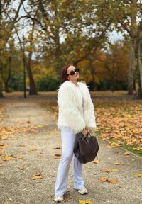 Biely kožúšok z umelého kožúcha, sivé nohavice a béžové topánky. Tmavá kabelka držaná v páse. Pozadie ukazuje stromy so zimnými listami.