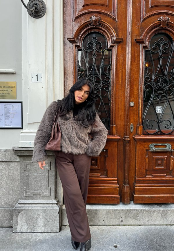Ruske faux fur jakna, crni turtleneck, smeđe široke hlače i mala smeđa torbica, stojeći pored ukrašenih drvenih vrata s metalnim detaljima.