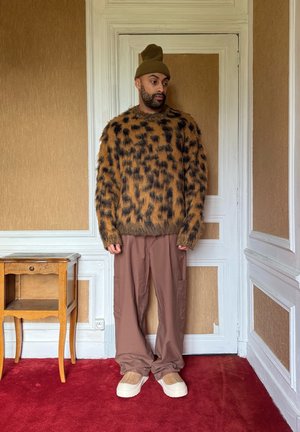 Jersey gris con patrón marrón y negro, pantalones cargo beige y zapatos de tacón crema. Un gorro verde completa el conjunto. Fondo de alfombra roja.