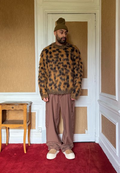 Jersey gris con patrón marrón y negro, pantalones cargo beige y zapatos de tacón crema. Un gorro verde completa el conjunto. Fondo de alfombra roja.