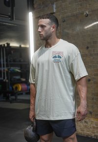 T-shirt oversized bege claro com uma estampa gráfica, usado com calções escuros. A pessoa segura um kettlebell preto num ambiente de ginásio.