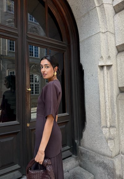 Una mujer lleva un vestido marrón ajustado con mangas cortas, acompañado de un bolso de mano de mimbre marrón. El fondo presenta una puerta de madera arqueada.