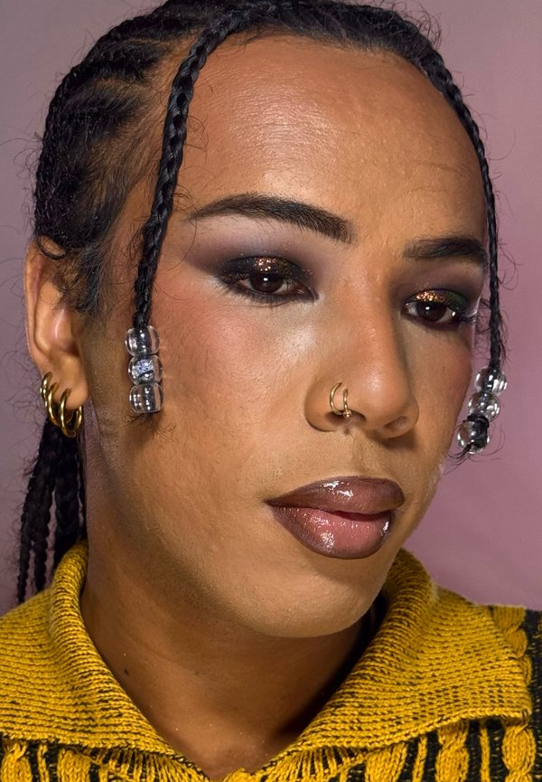 Personne avec des cheveux tressés ornés de perles transparentes, des boucles d'oreilles créoles en or, un anneau nasal en or, portant un maquillage des yeux foncé et un pull en maille jaune.