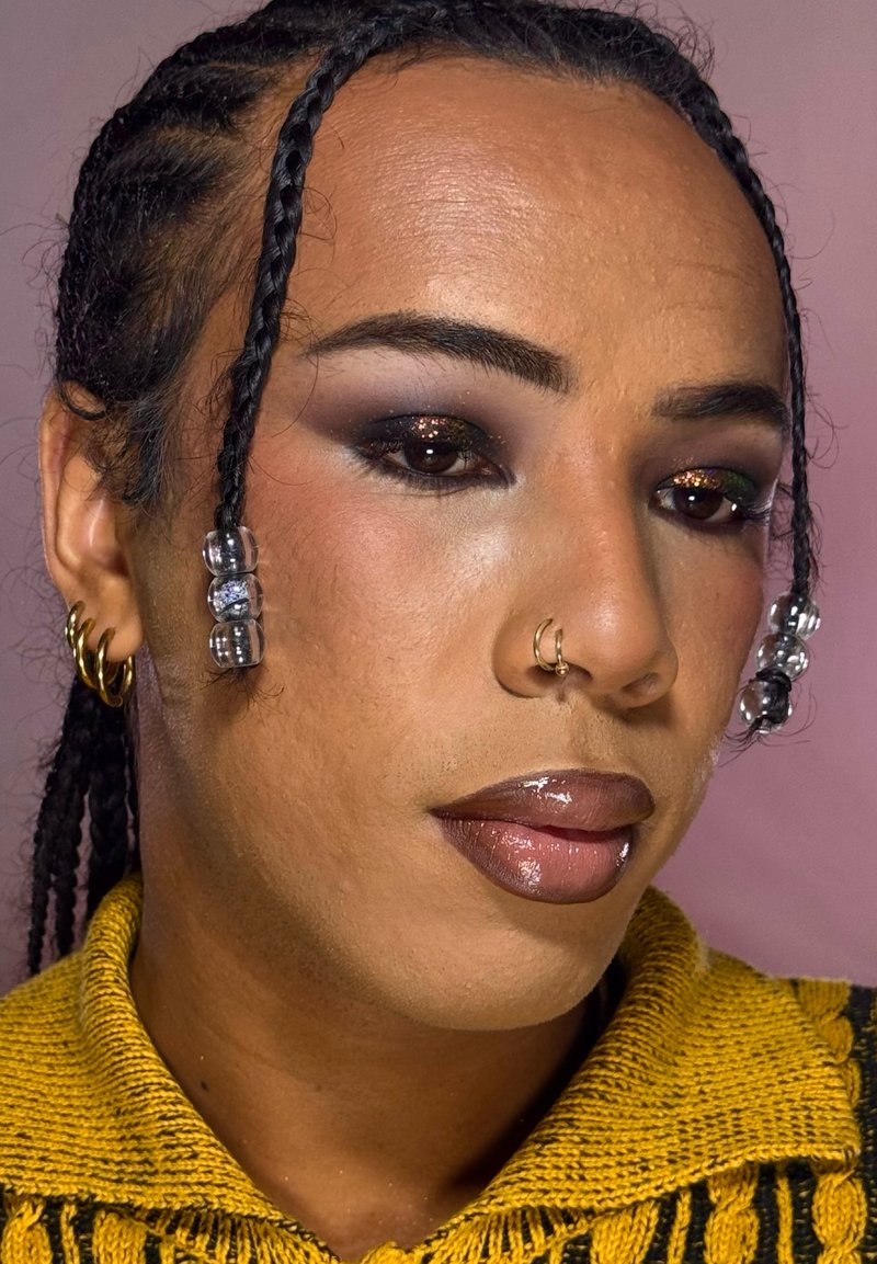 Person mit geflochtenem Haar, geschmückt mit klaren Perlen, goldenen Creolen, einem goldenen Nasenring, trägt dunkles Augen-Make-up und einen gelben Strickpullover.