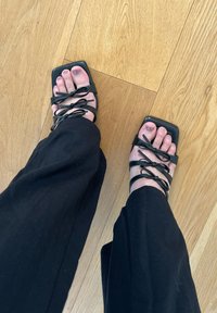 Sandales à talons hauts noires à lanières avec un design lacé, bout carré et texture lisse, portées avec un pantalon large noir sur un sol en bois.
