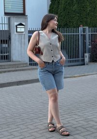 Lys beige, ermeløs vest med knapper, kombinert med lyseblå denimshorts. Brune sandaler og en liten brun håndveske fullfører antrekket.